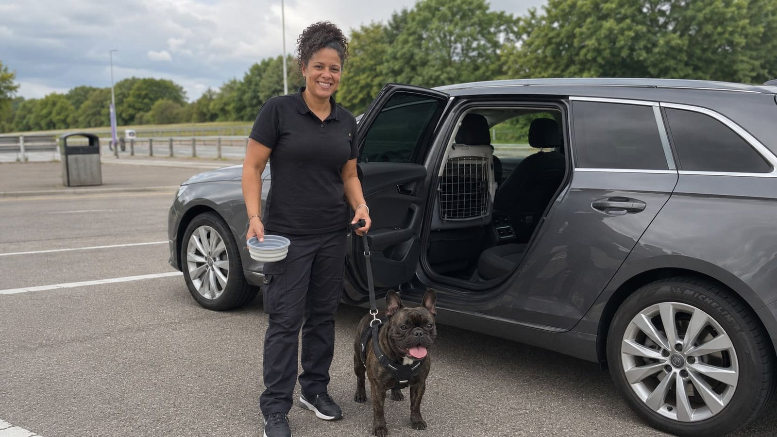 Pet chauffeur stands beside a harnessed dog and open vehicle.